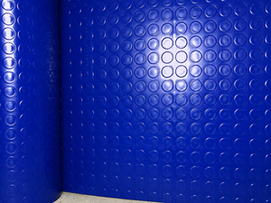 A close-up of a bright blue textured surface with a repeating circular raised pattern, shown as a wall panel or rolled material edge against a neutral floor.