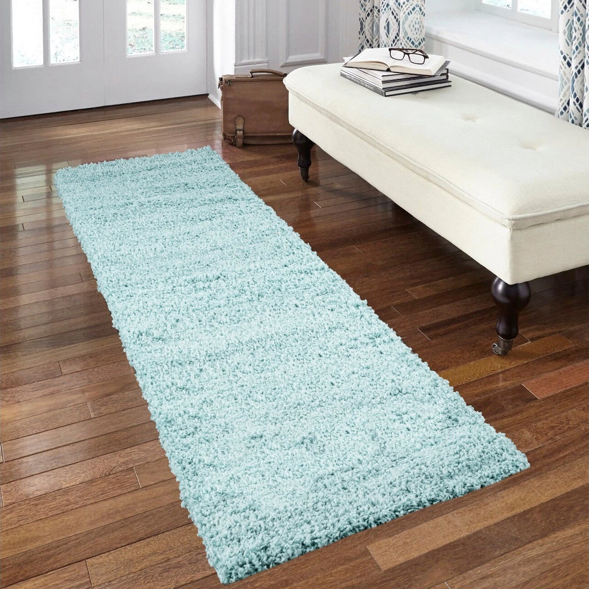 A long, light blue shag runner rug laid out on a dark wood floor in a bright room, with a cream tufted bench and white door visible in the background.
