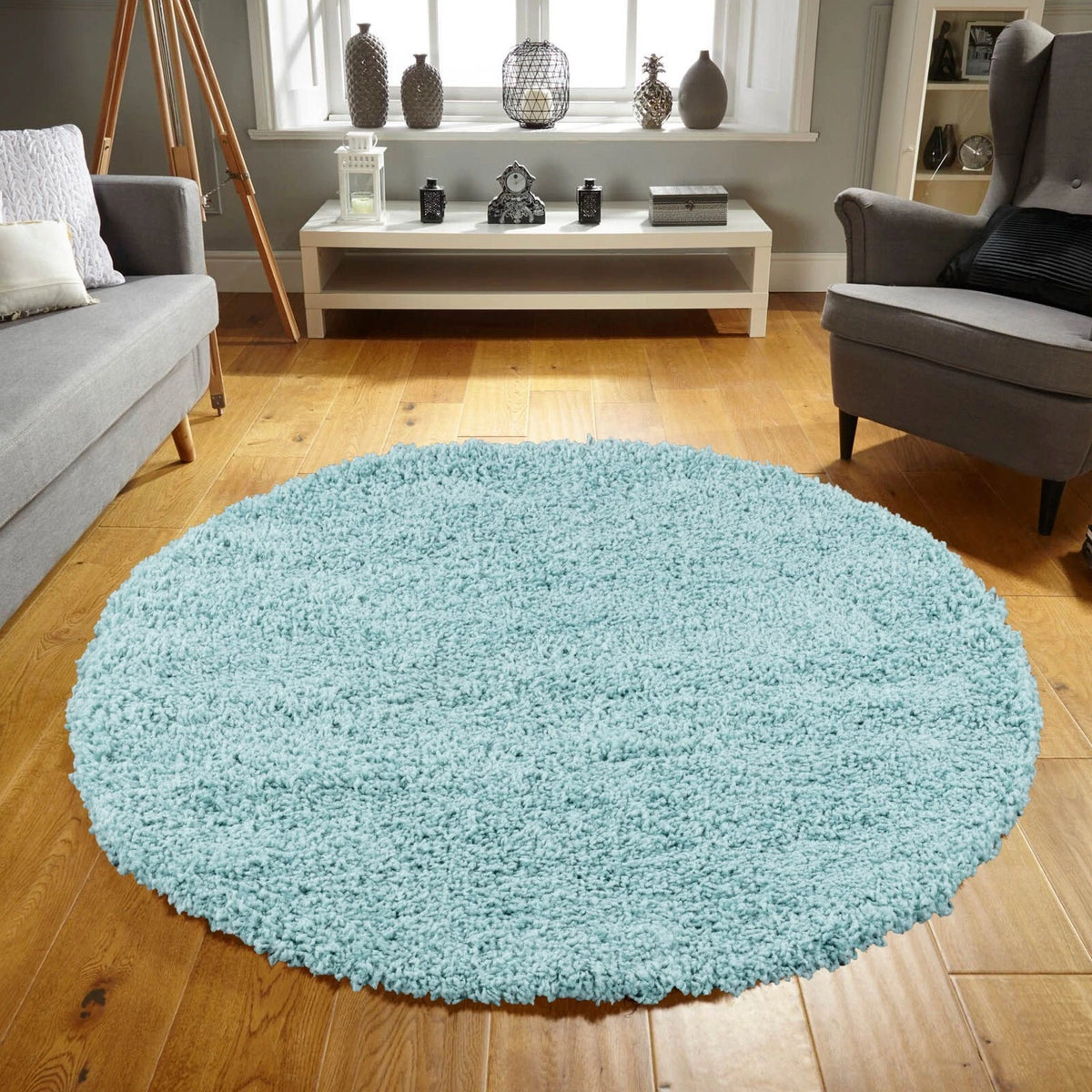 A round, light blue shag rug placed on a wooden floor in a modern living room setting with a gray sofa, armchair, and low console table.