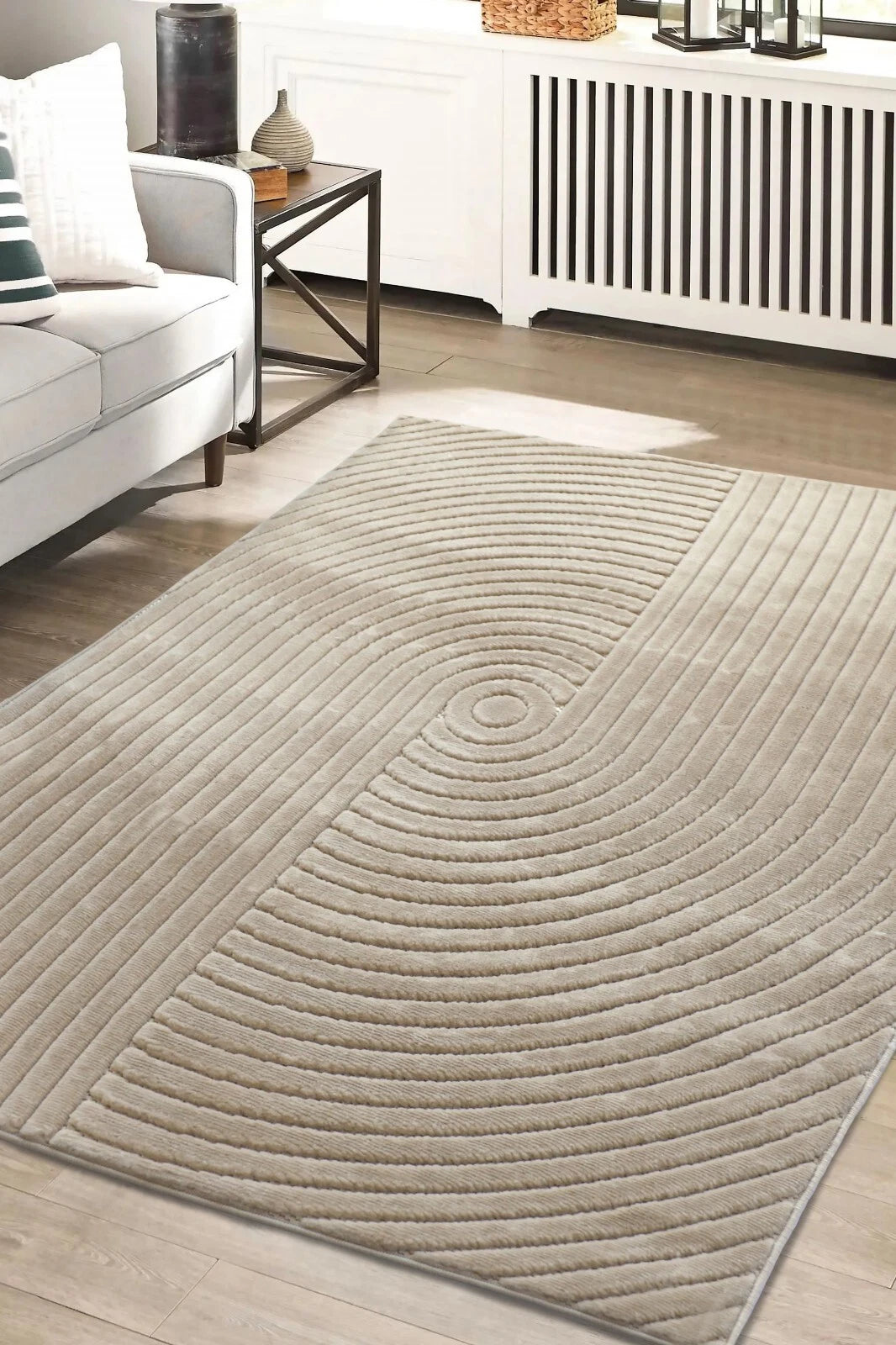 A rectangular area rug with a textured concentric circle pattern in a light beige/taupe color, shown on a light wood floor in a modern living room setting.