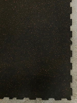 Black interlocking foam exercise mat with a textured surface and small orange flecks, shown partially cut off on a light gray floor.