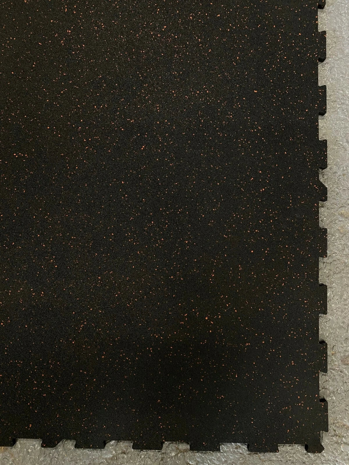 Black interlocking foam exercise mat with a textured surface and small orange flecks, shown partially cut off on a light gray floor.