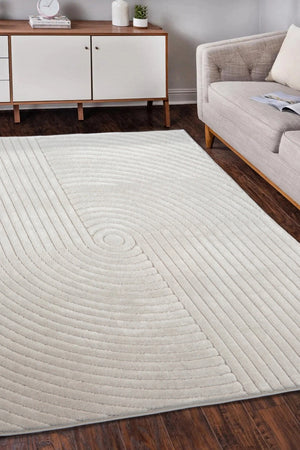 A large rectangular area rug in a warm off-white or cream tone with a raised concentric circular and radiating linear texture, shown on a dark wood floor in a living room setting next to a light gray sofa.