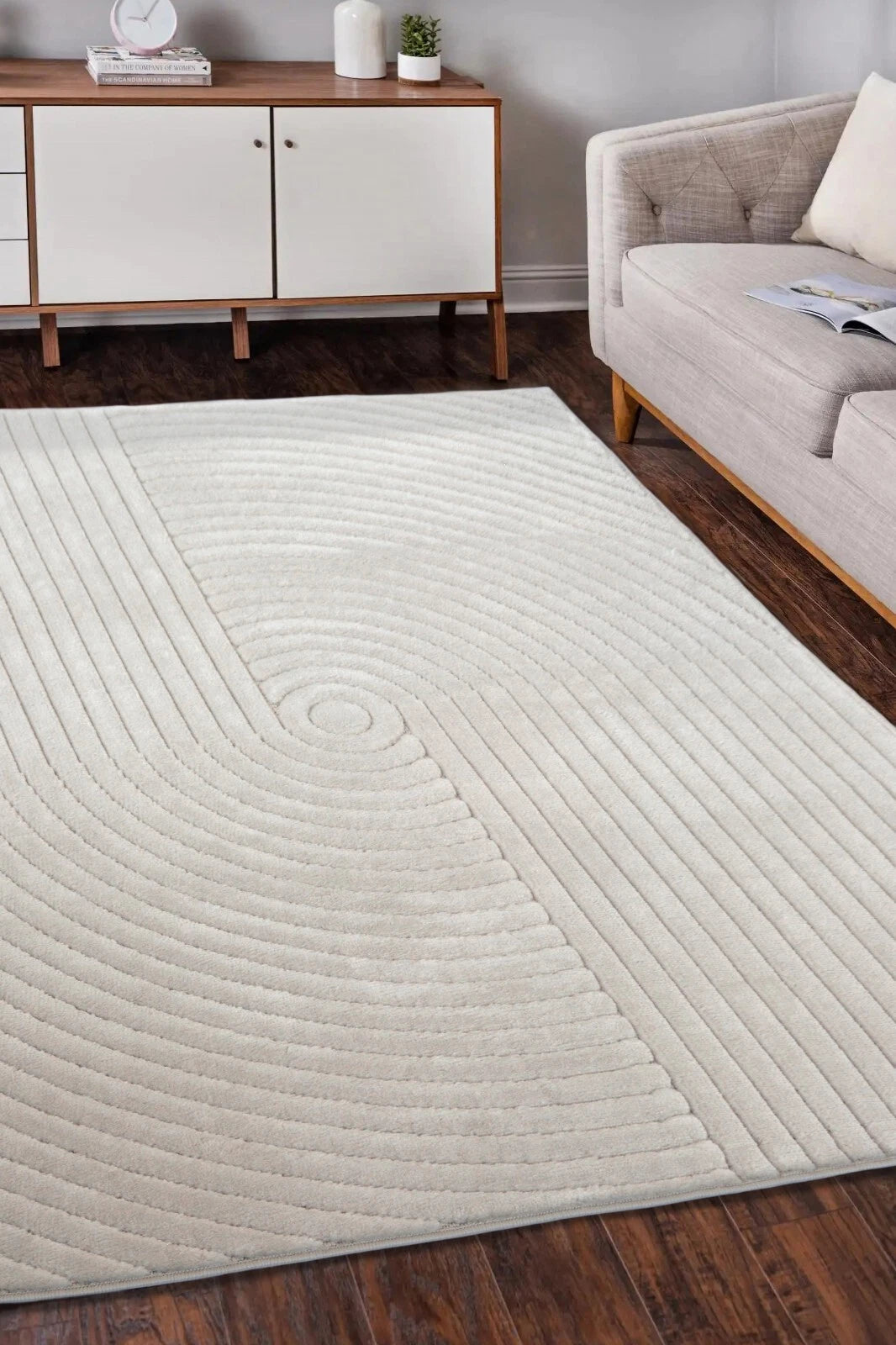 A large rectangular area rug in a warm off-white or cream tone with a raised concentric circular and radiating linear texture, shown on a dark wood floor in a living room setting next to a light gray sofa.