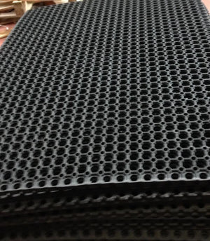 A close-up of a black plastic grid mat with a repeating honeycomb-style circular hole pattern, stacked on top of another mat.