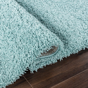 A close-up of a plush, high-pile shag rug in a light minty aqua blue color, shown folded over to reveal its underside and texture, resting on a dark brown wood floor.