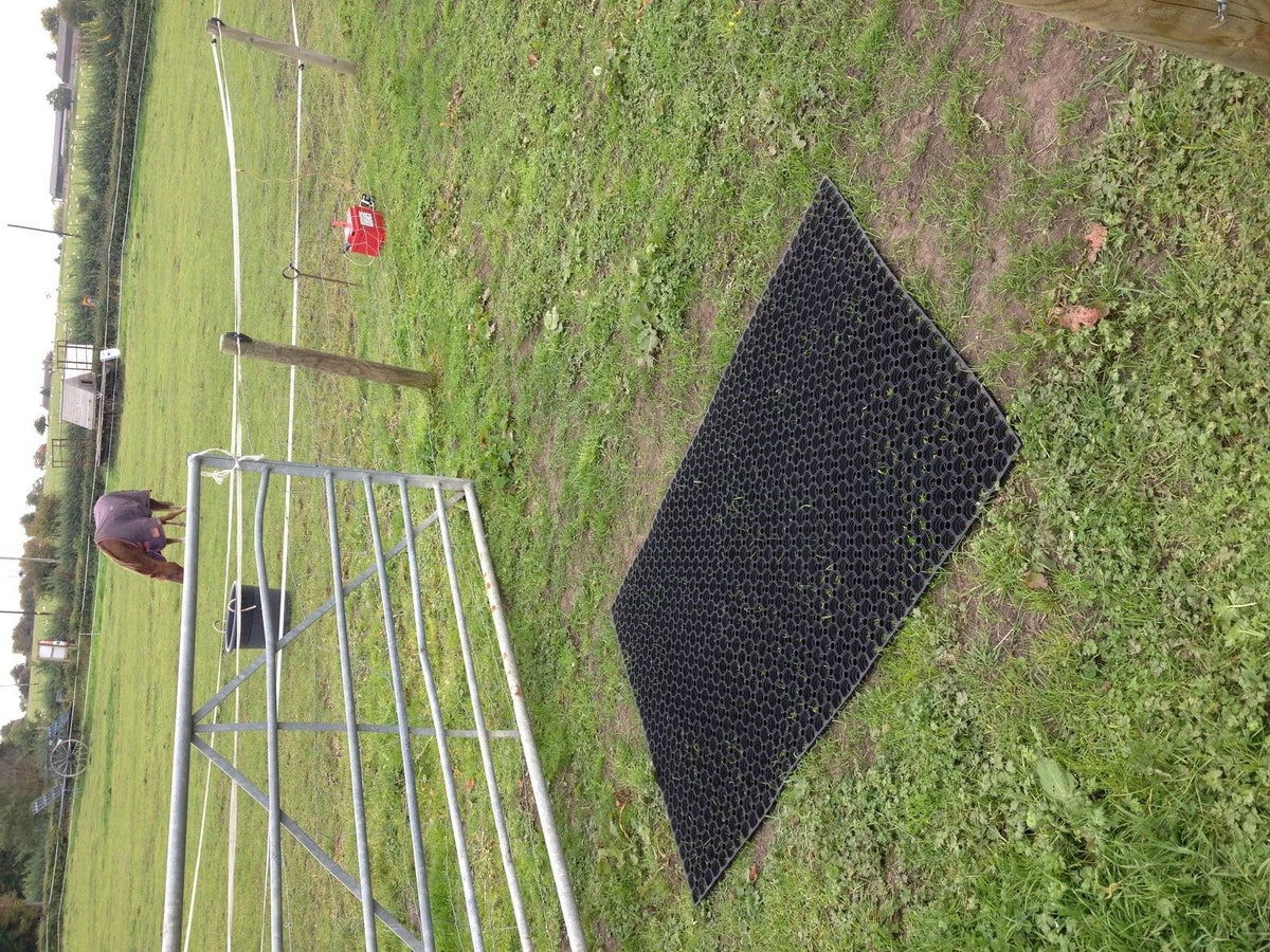 A black rubber horse gate mat with a honeycomb pattern, shown on a grassy outdoor paddock next to a metal gate.