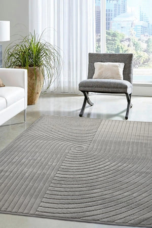 A large gray area rug with a textured concentric circle pattern, shown in a modern living room setting with a gray armchair, white sofa, and potted plant.