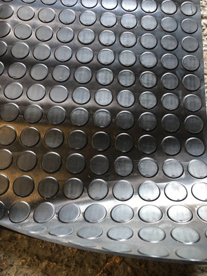 A close-up of a dark gray metallic sheet with a repeating pattern of raised circular domes, photographed against a rough concrete-like surface.
