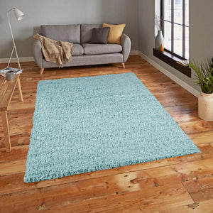 A light blue shag area rug displayed in a modern living room setting with a gray sofa, wooden floor, and a window.