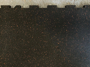 Close-up of a black interlocking rubber gym floor mat with small orange-brown flecks, showing the tongue-and-groove edge against a light textured surface.