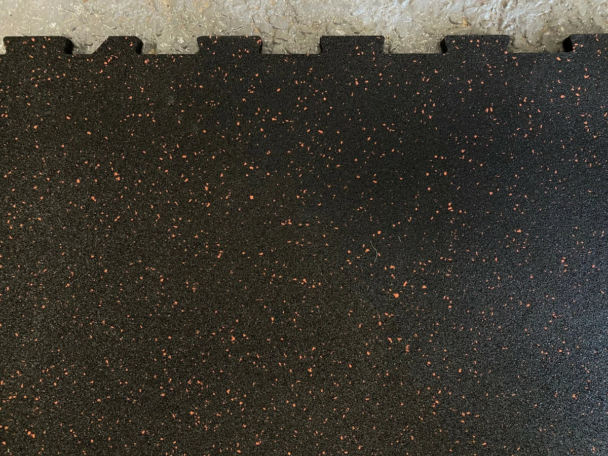 Close-up of a black interlocking rubber gym floor mat with small orange-brown flecks, showing the tongue-and-groove edge against a light textured surface.