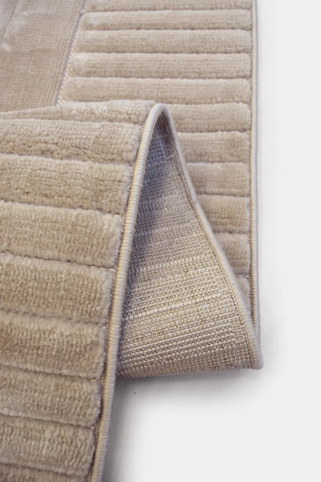 Close-up of a beige textured rug with a ribbed plush top and a woven underside, folded over on a white surface.
