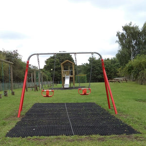 Black heavy-duty rubber grass mats installed on lawn ground underneath a red metal swing set in an outdoor playground park.