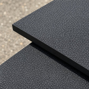 Two textured black foam mats stacked diagonally, showing a rough, pebbled surface and thick edge, with a blurred gray gravel background.