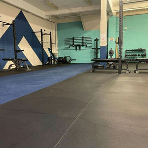 A wide perspective view of a fitted commercial gym floor covered in non-slip black rubber floor tiles under workout benches and weight training equipment.