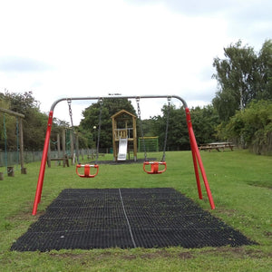 A children's playground swing set with two red swing seats and a black rubber mat foundation, set on a grassy lawn with a wooden playhouse and trees in the background.
