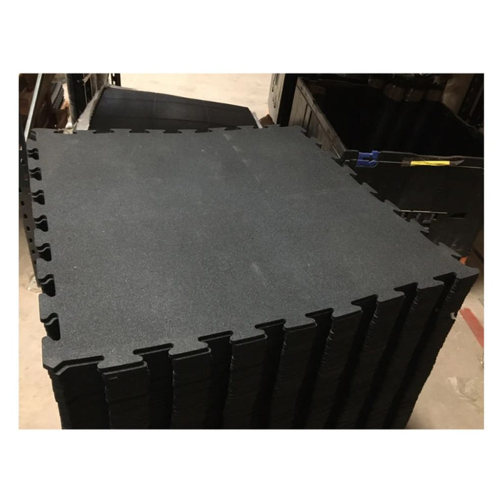 A stack of black interlocking foam floor tiles with a textured surface, photographed in what appears to be a warehouse or storage area.