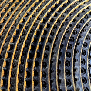 Close-up of a black textured material with a repeating pattern of circular cutouts and raised gold-orange stitched or printed lines running parallel to it.