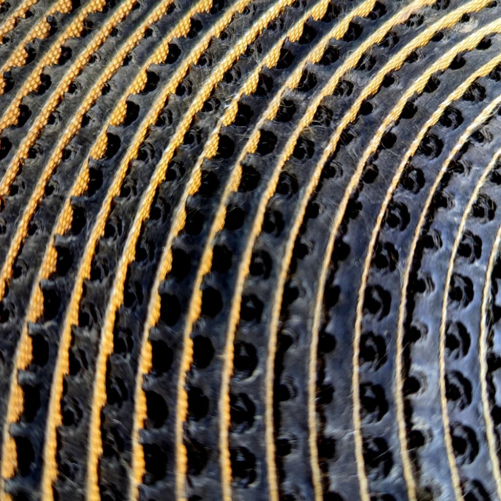 Close-up of a black textured material with a repeating pattern of circular cutouts and raised gold-orange stitched or printed lines running parallel to it.